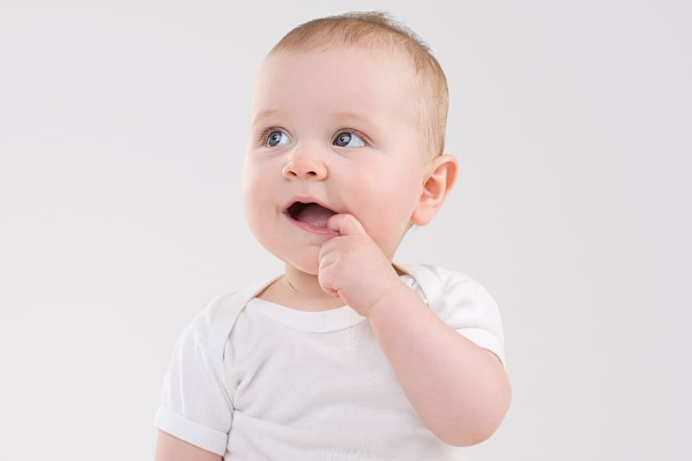 Smiling baby in a white outfit with a finger in their mouth, looking slightly to the side against a plain background